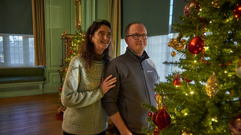 A man and woman stand in the Drawing room looking at the Christmas tree. The room is large with a big window behind them and wooden floors. The walls of the room are light green.